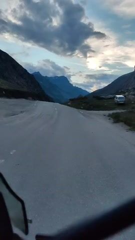 A first-person perspective video captures a paved road winding through a mountain valley. The road is grey asphalt with white lane markings. Steep, dark mountains flank the road on both sides. The sky above is filled with dramatic, dark clouds, with a sliver of bright sunlight breaking through in the distance. The sun appears to be setting or rising. A black mirror housing is visible in the lower left corner of the frame, suggesting the footage is from a vehicle. A white van is parked on the right side of the road in the distance. The overall impression is one of vast, rugged natural beauty and a sense of journey.