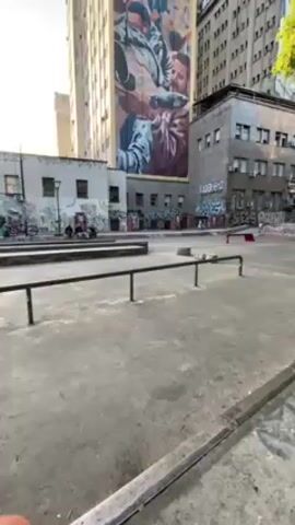 The video shows a skateboarder attempting a trick at an urban skate park. The skateboarder, wearing a black t-shirt and blue jeans, rides towards a metal rail.  He attempts to jump onto the rail with his skateboard, but fails, landing on the ground.  The skate park has graffiti on some of the walls and the ground is concrete. In the background, there are tall buildings, and the sky is blue with some clouds. Other people are present in the park, either sitting or standing around the perimeter.