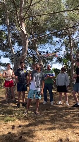 In an outdoor scene set amongst sparse trees with fallen pine cones covering the ground, a group of seven individuals is seen. The man in the center, wearing a gray and white t-shirt and blue patterned shorts, is holding a cell phone and appears to be speaking. The others are arrayed around him, listening. To his left, a man in a tank top and red shorts stands with his arms crossed. Next to him is a man in a graphic t-shirt and dark shorts, also looking at the speaker. To the right of the speaker is a man in a green and blue striped shirt and jeans, holding a notebook. Next to him is a man in a gray hoodie and dark shorts. Further to the right, a man in a black t-shirt and dark shorts is also watching. The sky is visible through the trees, indicating a bright day. The overall impression is a casual, informal gathering.
