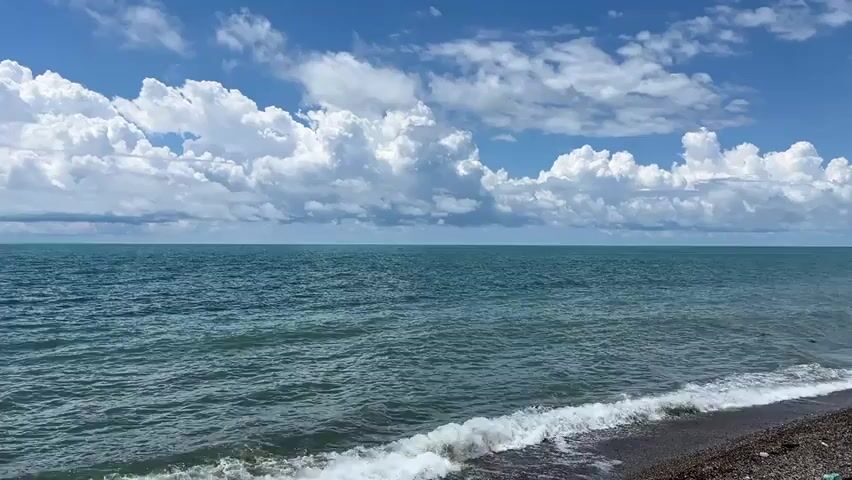 A wide shot of a calm sea under a partly cloudy sky. The water is a deep turquoise color and there are gentle waves lapping at a pebble beach in the foreground. The sky is a vibrant blue with large, fluffy white clouds scattered across it. The horizon is clearly visible, separating the sea from the sky. The scene appears peaceful and serene.