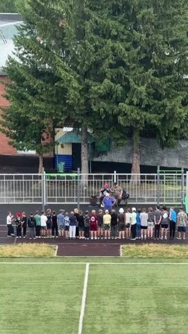 A group of children and adults are gathered on a grassy field in front of a fence. The children are standing in a line, facing a man in a blue shirt and shorts who appears to be holding a white ball. The adults are standing behind the children. There are large evergreen trees behind the fence, and a brick building to the left. The field appears to be a sports field, with white lines marking boundaries. The sky is overcast.