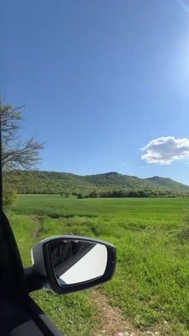 The video starts with a view from a car window, showing a bright, sunny day with a green field and a small mountain in the background. The camera pans slightly to reveal the car's side mirror and then moves inside the car. There are four people in the car: two women in the front, and a man and another woman in the back. The camera focuses briefly on the man in the back, who looks at the camera. The camera then pans back to the window, showing the green field and mountain again.