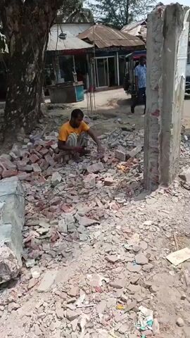 Here's a plain text description of the video:

The video shows two men working outside on a demolition or construction site of some kind.  

In the first scene, a man in a yellow shirt is squatting and appears to be using a hammer or chisel to break up brick debris. He's surrounded by broken bricks and gravel. In the background is a building and a parked autorickshaw.

In the second scene, a different man, wearing a maroon shirt and patterned pants, is standing behind a small wooden table, using a sledgehammer on a pole. There is brick debris on the ground. In the background are other buildings, a person walking by, and an autorickshaw.