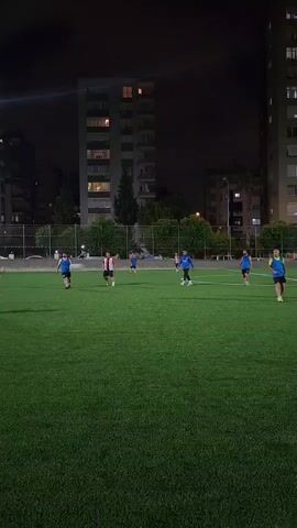 A group of men are playing soccer on a green turf field at night. The field is lit by stadium lights. In the background, there are tall apartment buildings and trees. The men are wearing various colored shirts and shorts. One man in a red and white striped shirt is dribbling the ball. Another man in a blue shirt is running towards him. The other players are spread out on the field. The overall scene depicts a casual game of soccer.