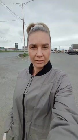 A woman stands in front of a camera, speaking. She is wearing a grey bomber jacket with black trim and her blonde hair is pulled back in a ponytail. She appears to be outdoors, in what looks like a parking lot or industrial area. In the background, there are several cars, some buildings, and a cloudy sky. The woman is speaking directly to the camera, and her expression is serious. She gestures with her hands as she speaks. The camera is held at chest level, giving a first-person perspective.