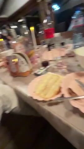 The video shows a long table with several pink plates, some with food on them, and various glasses and bottles. There's a small remote-like device on the table near a napkin holder. The lighting seems indoor and somewhat dim. The camera is shaky and moves around, suggesting it's handheld. The focus is soft, blurring much of the scene.