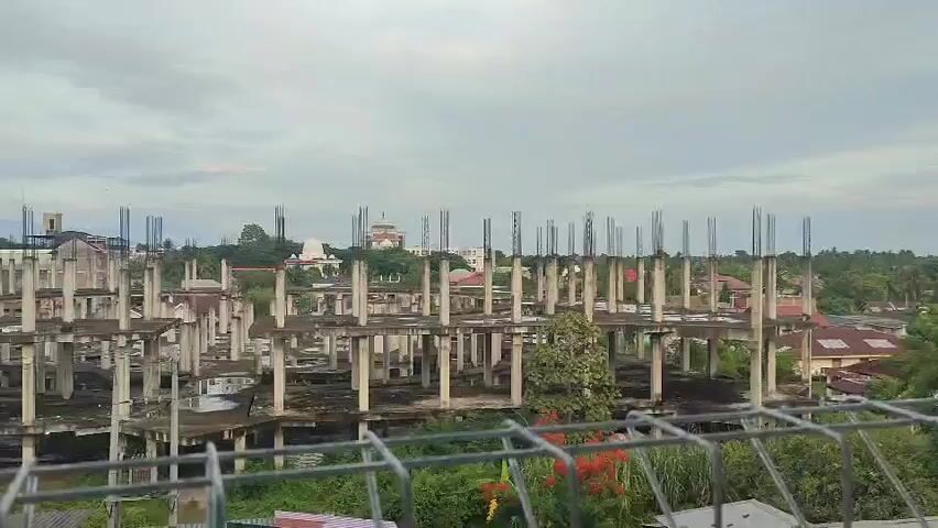 The video shows an unfinished construction site with concrete pillars and floors of multiple buildings. Some of the structures are partially complete with exposed rebar. In the background, there are trees, other buildings, and a slightly cloudy sky. In the foreground, there is a metal fence. The scene is static with no apparent movement or activity.