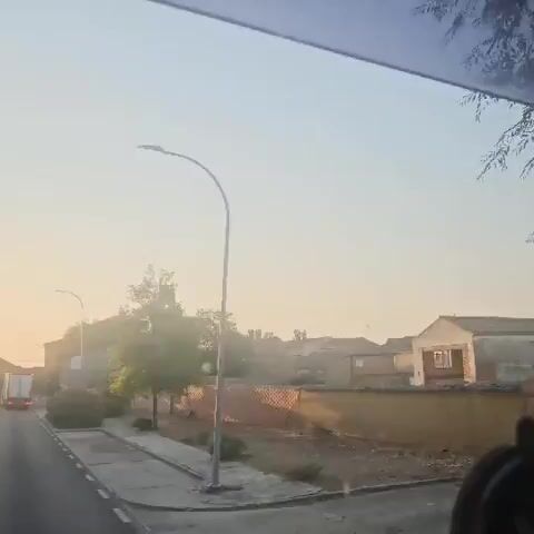 The video captures a view from a vehicle, likely a bus, moving along a road during what appears to be sunrise or sunset due to the warm, soft lighting. The sky is a pale, hazy blue, transitioning to yellow near the horizon. On the right side of the road, there are houses with terracotta roofs and stone walls. A prominent feature is an old stone bell tower with a cross on top, partially obscured by trees. A bus stop sign with the emblem of "Junta de Castilla y León" and the word "BUS" is visible in the foreground. On the left side of the road, there's a sidewalk and some greenery, including a lone tree. A white truck is seen driving away from the camera on the left side of the road. The overall scene suggests a rural or semi-rural setting.