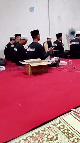The video shows a group of men sitting on a red carpeted floor in a room with white walls. Most of the men are wearing black t-shirts with white lettering and black skullcaps. Some are holding musical instruments, including what appears to be a frame drum. One man is actively playing the drum. There is a cardboard box in the foreground. A fan is visible on the right side of the room. The overall atmosphere appears to be a musical performance or rehearsal.