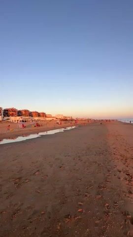 A vertical, low-angle drone shot glides over a wide, sandy beach towards the ocean at dusk. The sky is a clear gradient of blue fading to a soft orange near the horizon. The sand is packed with footprints and scattered debris. Along the left side of the frame, a row of buildings with red roofs and white awnings faces the beach, and colorful umbrellas and beach chairs are visible. A small body of water, possibly a canal or stream, separates the buildings from the main beach. People are scattered across the beach and in the water, some walking, others relaxing. The ocean waves gently lap the shore. In the foreground, a single, small, white and black object rests on the sand.