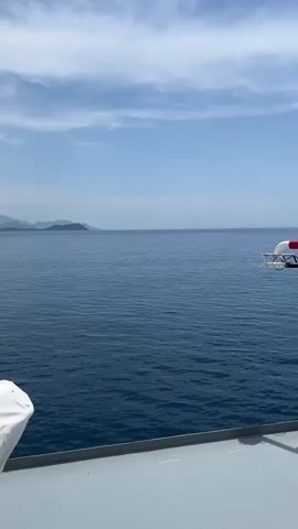 The video opens with a view of the ocean and distant islands under a cloudy sky. The camera then pans to show a covered lifeboat on the deck of a boat. The view shifts to show the deck of the boat, with rows of white seats, and then a wider view of the deck with several people visible. One person on the left appears to be tethered to a rope. Another person in the center is leaning over a railing. The video ends with a view of the ocean and islands.