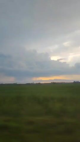A video shows a blurry green field under a cloudy sky. The horizon is visible with a strip of orange light peaking through the clouds. There are some faint outlines of buildings or structures in the distance. The camera appears to be moving, causing the blur in the foreground.