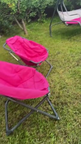 The media file depicts a backyard setting in Тмз Снт, Russia. The scene features a swing chair with pink and white cushions, two pink folding chairs, and a wooden gazebo with white curtains. An inflatable pink flamingo float is visible inside the gazebo. The lawn is green and well-maintained, with several green bushes and trees surrounding the area. A paved pathway made of rectangular stones is also present.

The overall impression is that of a relaxed and inviting outdoor space, likely prepared for leisure or relaxation. The presence of the swing chair, folding chairs, and the flamingo float suggests activities such as lounging, sunbathing, or possibly swimming if a pool is nearby (though not visible in the frame).

The lighting suggests it is late afternoon or early evening, with the sun casting a warm glow on the trees and gazebo. The sky is partially visible and appears to be a clear blue, indicating pleasant weather. There is no visible text in the media file. The atmosphere conveyed is peaceful and serene, suggesting a tranquil environment.