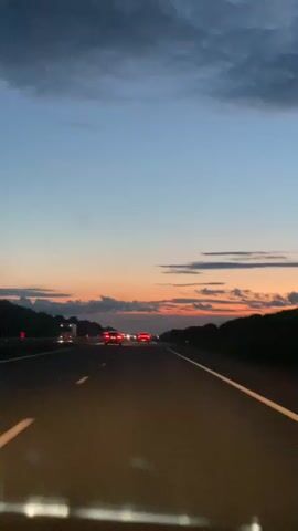 The video shows a first-person perspective of a car traveling on a highway at dusk. The sky transitions from a pale blue at the top to orange and pink hues near the horizon, indicating the sun is setting. Dark clouds are present in the upper portion of the sky. The highway has clear lane markings, with a white line visible on the right side of the road. Other vehicles are visible on the highway, their headlights and taillights illuminated. Trees or dense foliage line the sides of the highway. The overall impression is of a peaceful and scenic drive as nightfall approaches.