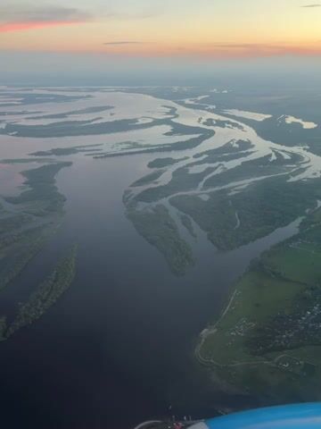This aerial footage captures a vast, meandering river system with numerous islands covered in lush green trees. The water is a deep, dark blue, contrasting with the lighter reflections on its surface and the lighter hues of the land. The landscape is predominantly natural, with extensive forested areas and the intricate network of river channels and islands.

In the distance, a hazy horizon suggests a broad, flat landscape. The sky above is painted with the soft colors of dawn or dusk, featuring pastel shades of pink, orange, and pale blue. This warm, soft lighting indicates it is either early morning or late evening.

On the right side of the frame, the riverbank is visible, with patches of green fields and a cluster of small buildings, suggesting a rural settlement. A road winds along the riverbank, connecting the settlement to the surrounding area. The overall impression is one of natural beauty and tranquility, emphasizing the scale and majesty of the river and its surroundings. The camera appears to be mounted on a vehicle or drone, indicated by a sliver of a blue object at the very bottom of the frame, possibly part of the craft. There is no visible text within the footage.