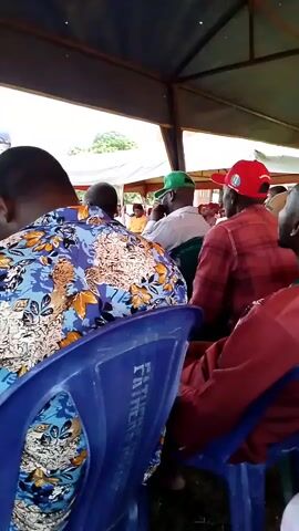 The video shows an overhead view of a group of men sitting in chairs under a tent or canopy. The chairs are blue and appear to be plastic. The men are mostly facing away from the camera. Some of them are wearing hats. The tent has a brown metal frame and a light-colored fabric roof. The ground beneath the tent appears to be covered in grass. In the background, there is some greenery and what looks like a white tent or structure. The lighting suggests it is daytime.