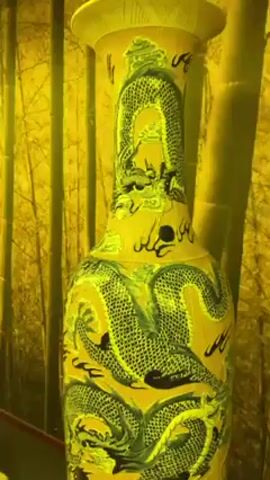 The video starts with a close-up of a yellow vase decorated with dragons. The camera then pans out to reveal a long hallway. The hallway has yellow walls with a design reminiscent of bamboo. It has a wooden floor and a white railing. Above the hallway, lights are hung. People are walking in the hallway.