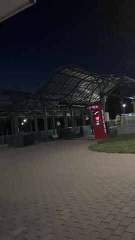 A public space at night, possibly a park or a bus stop, with a covered waiting area. A red vending machine is visible. A few people are present, some sitting and some walking. The ground is paved with interlocking bricks.