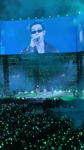The video shows a live concert scene with a large crowd holding up green lights in a darkened stadium. A performance is taking place on a circular stage, where several people are visible in various outfits. Above the stage is a massive screen displaying a man wearing sunglasses and holding a microphone. He appears to be performing as well, likely projected for the audience. The stage is surrounded by metal rigging and illuminated by green lighting and spotlights. The overall ambiance indicates a vibrant and energetic event.