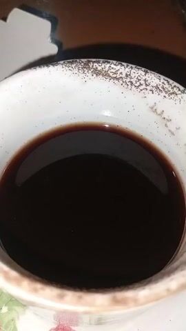 A close-up shot shows the dark liquid in a white ceramic cup with speckled brown edges. Some faint floral designs are visible on the lower portion of the cup and saucer. The liquid is still and reflects light from above.