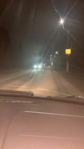 The video shows a nighttime view from inside a car driving down a road.  The road is lit by streetlights, and other cars can be seen in the distance. There is a road sign visible on the right. The dashboard of the car is visible.