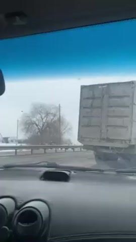 The video is taken from the inside of a car, focusing on the road ahead. The weather appears snowy or overcast. A large, gray truck with a boxy container-like back is directly in front of the camera car.  Other cars, a black sedan and a white SUV, can be seen in the adjacent lane. A bare tree stands to the left of the truck, beyond a metal guardrail. Utility poles and wires are visible in the background. The dashboard of the camera car is in the foreground. The windshield wipers appear to be on.