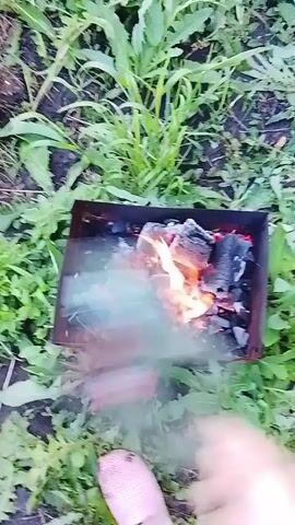 A video shows a close-up view of burning charcoal in a metal grill on the ground, surrounded by green grass. The camera is angled downwards, and a person's foot is visible in the bottom left corner of the frame. Smoke rises from the grill. The video captures the initial moments of the fire.