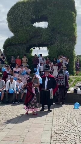 The video shows a group of people gathered in a public area, likely a park or plaza, with a unique backdrop of large, bush-shaped letters. Most of the crowd is seated on tiered concrete steps. The people wear a mix of casual clothing and traditional attire. A young girl in a floral dress and a red shawl dances and waves an Argentine flag, standing in front of a man in traditional clothing and a wide-brimmed hat, who appears to be watching her. The crowd holds blue balloons. The area is paved with brick. The sky is overcast.