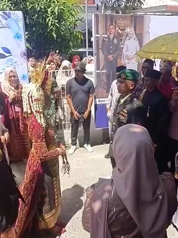 A group of people are gathered outdoors, possibly for a wedding or a formal event. In the foreground, two individuals in elaborate traditional attire, one appearing to be a bride with a tall, ornate headdress, are facing each other. A man in a black t-shirt and baseball cap stands behind the bride. To the right of the bride, a man in a military uniform, wearing a green beret, is facing towards the other individuals. Behind him, a large banner displays wedding portraits of a couple. Several other people, some wearing headscarves and others in formal wear, are observing the scene. A yellow umbrella is held above some of the individuals. The environment appears to be paved with some foliage visible in the background.