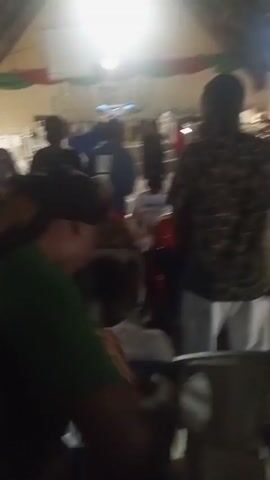 The video appears to be filmed inside a room, possibly a church or event hall, with many people present. The lighting is dim, and the camera is unsteady, causing a blurry effect. In the foreground, a person wearing a green shirt is visible, with their back to the camera. In the background, multiple individuals are standing and seated, some engaged in conversation or movement. The room has a high ceiling, and decorative banners are visible on the walls. There are chairs arranged in rows, and a stage or elevated platform is present at the front of the room. The overall atmosphere suggests a gathering or event is taking place.