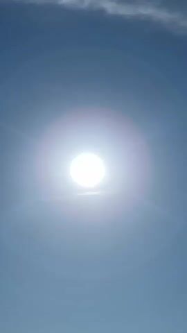 A bright sun is visible in the center of the frame, shining through a clear blue sky with a wispy cloud in the upper left corner. A person wearing a dark shirt and a baseball cap is walking in the lower left quadrant of the frame. In the lower right quadrant, part of an orange object, possibly a tent or blanket, is visible. A body of water is visible in the background behind the person.