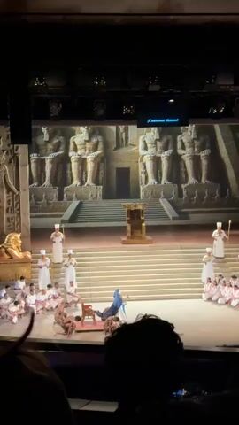 Here is a description of the scene in the video.
The video shows a stage performance depicting an ancient Egyptian scene. In the background is a large screen displaying a temple-like structure with four statues. In the foreground, actors in white robes stand or kneel on tiered steps, some holding staffs. A group of bare-chested actors are bowed down on the floor. A golden throne stands center stage. A figure in a blue robe is also present. The words "Gloria al rey!" are projected on the screen. The video is taken from the audience, with the heads of audience members visible at the bottom.