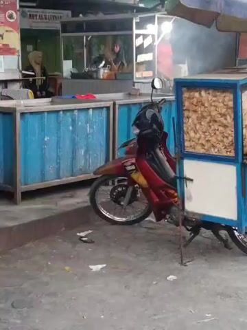 A red and yellow motorcycle is parked in front of a food stall. The stall has a blue metal counter and a sign that reads "BMT AL-KAUTSAR". There are several people working at the stall, preparing food. Smoke is coming from the stall, indicating that cooking is in progress. The ground is paved with concrete.