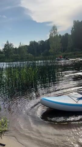 A man and a woman are sitting on a paddleboard in the water. The paddleboard is blue and white. The man is wearing swim trunks, and the woman is wearing a swimsuit. They are both looking forward. The water is calm, and there are trees in the background. The sky is blue with some clouds. The sun is shining. There is a lot of green vegetation in the foreground.