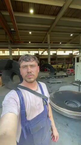 This media file showcases a man in blue overalls and a light-colored t-shirt, presumably a worker, posing for a selfie. He is inside a large industrial building with exposed beams and skylights, suggesting it's daytime. The man appears to have dirt or grease on his face and clothes, indicating a hands-on job. In the background, various metal objects, possibly parts of machinery or large circular components, are visible, along with some equipment like a forklift.

The scene then shifts to an outdoor industrial or scrapyard area, with a clear sky and some clouds, suggesting fair weather. This outdoor portion of the media displays piles of scrap metal, discarded items, and industrial machinery. In the distance, rolling hills and mountains characteristic of Tomba, Montenegro, are clearly visible under a bright sky. The contrast between the indoor industrial setting and the scenic mountainous landscape of Montenegro is striking. The outdoor shots reveal a mix of old and new structures, with some operational equipment, such as an orange and gray industrial unit with a fan, and a red tractor-like machine. The overall impression is of a working industrial environment set against a beautiful natural backdrop.