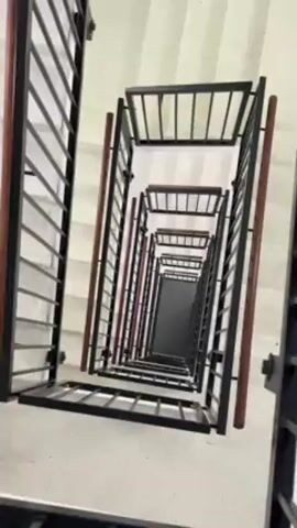 A high-angle shot looks down a multi-story staircase. The stairs are white, and the railings are black metal with wood accents. The camera slowly rotates, revealing more of the staircase as it descends. A person's foot briefly appears at the bottom of the frame. The environment appears to be an indoor stairwell, possibly in a residential or commercial building.