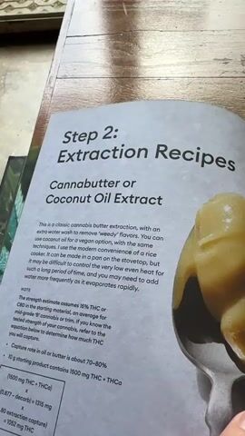 The video shows a close-up of a recipe book open to a page titled "Step 2: Extraction Recipes Cannabutter or Coconut Oil Extract". The page contains text describing methods for making cannabis extracts and a recipe for cannabutter or coconut oil. To the right of the book, a spoonful of a yellow, creamy substance, likely cannabutter or coconut oil, is visible. The book is lying on a surface that appears to be a wooden table. The lighting suggests an indoor environment, possibly with natural light filtering from a window. There are no people or explicit actions captured in the video, only the static image of the recipe book and the extracted substance.