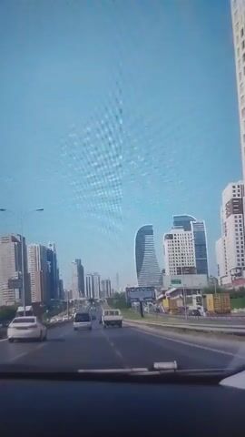The video shows a view from inside a car driving on a highway towards a city skyline. The sky is clear and blue. Several tall buildings are visible, including one with a unique curved shape. There are other vehicles on the road, including cars and a pickup truck. A billboard with the word "ONVO" can be seen on the side of the highway.