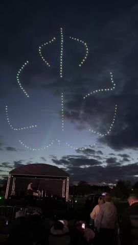 A crowd of people is watching a performance on a stage at night, with an array of drones in the sky forming a glowing, rounded T-shaped symbol. The stage is covered, and a single performer can be seen on stage. The crowd is mostly dark, but some people are holding up their phones to take photos or videos. The sky is a dark, cloudy blue, with a hint of light on the horizon. The drone formation is made up of small, bright lights, arranged to create a recognizable symbol. The scene appears to be part of an outdoor concert or event, taking place in a park or open space.