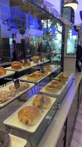 This video captures a close-up view of a bakery display case filled with an assortment of pastries. The display is brightly lit, with the primary illumination coming from overhead blue lights, casting a cool, modern ambiance. The pastries are artfully arranged on white platters, tiered for optimal visibility.

The subjects are primarily the baked goods themselves, including croissants, pain au chocolat, almond croissants, and other flaky, golden-brown pastries. In the background, through the glass and reflected in it, hints of the cafe setting can be observed. This includes coffee machines, a menu board with blue text, a small decorative plant, and what appears to be a person working behind the counter, though they are largely obscured. The reflections also show an outdoor scene with trees and daylight, suggesting the cafe is situated in an urban or suburban area.

The activity taking place is the presentation of these freshly baked goods, likely for sale. The time of day appears to be daytime, given the outdoor light visible through reflections. There are no clear indicators of weather, but the indoor setting is well-lit and inviting. The location cues from the prompt (Weehawken, United States of America) point to a specific context for this bakery.

Notable details include the various labels placed in front of some of the pastries, though the text on them is mostly illegible due to the angle and resolution. However, one label clearly reads "COLD BREW" near the coffee machines, and another visible label has the words "ALMOND CROISSANT". The overall impression is one of a well-stocked, contemporary bakery offering a tempting array of pastries. There's a sense of quiet efficiency, as customers are not visible, and the focus is on the appealing food items.