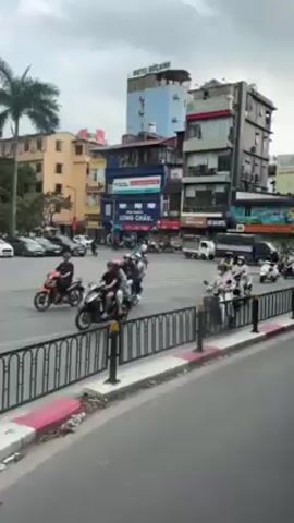 The video shows a busy street intersection in what appears to be a city in Vietnam. Numerous scooters and motorcycles are driving through the intersection, many carrying one or two passengers. There are a few cars and trucks as well. The buildings around the intersection are a mix of colors and heights, with advertisements and signs visible. A "no U-turn" sign is also present. The sky is overcast. The camera is positioned on the side of the road, behind a metal fence.