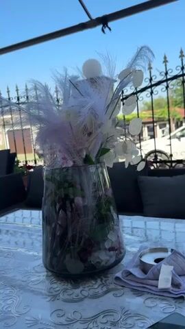 A clear, sunny day is depicted in this video, with the bright blue sky and warm lighting indicating it's likely midday. The scene is an outdoor seating area, possibly a patio or a cafe, given the presence of a table with a patterned tablecloth, comfortable-looking dark gray outdoor seating in the background, and a black metal fence separating the area from a street or property beyond.

The central focus is a tall, clear glass vase filled with an elaborate floral arrangement. The arrangement includes various elements, such as delicate white and pale pink feathers that fan out from the top, some round, pale discs that resemble lunaria or honesty plant seed pods, and hints of greenery and possibly darker flowers or petals towards the bottom. The vase itself is filled with water and contains more submerged foliage and decorative elements, adding depth and texture.

To the right of the vase, on the table, sits a metal ashtray on a folded light purple cloth. The presence of the ashtray suggests a relaxed, possibly social setting where smoking might be permitted. The overall mood appears peaceful and serene, enhanced by the gentle swaying of the feathery elements in the arrangement, possibly due to a light breeze. The video captures a static shot of this table setting, allowing the viewer to appreciate the details of the arrangement and the outdoor environment. No people are visible in the frame, and no visible text is present.