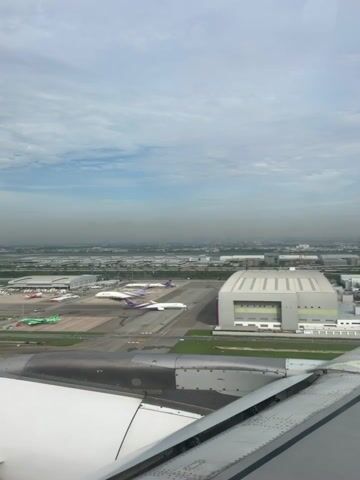 The video shows an aerial view of an airport tarmac with multiple airplanes on the ground. In the foreground, part of an airplane wing is visible, with its flap and engine housing. The tarmac features several aircraft, including white and purple planes that appear to be part of the same airline, and a few green planes. In the background, there are airport buildings, hangars, and a distant cityscape obscured by haze. The sky is partly cloudy. The camera appears to be mounted inside an airplane, looking out a window, as the video captures the subtle movements of an aircraft in motion, possibly taxiing or preparing for takeoff. There are no people visible in the frame.