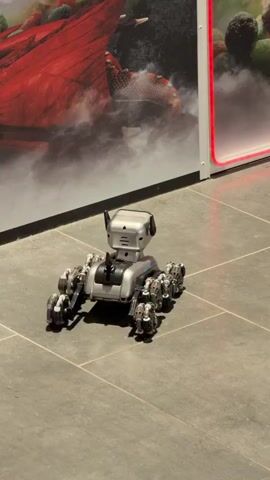 A small, robotic, rover-like toy sits motionless on a gray tiled floor. It has a silver and black body with multiple small wheels on each side. The "head" of the robot has two blue lights that resemble eyes, which illuminate as the video progresses. Behind the robot is a wall with two posters, one with a red creature and smoke, and the other with an image of berries. A strip of light borders the second poster. There is no visible movement or sound beyond the initial still image and subsequent lighting of the robot's eyes.