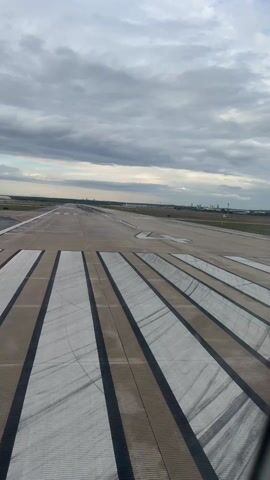 The video shows an aerial view of an airport runway on an overcast day. The runway is made of concrete, and is marked with white and black stripes. In the distance, other runways and airport infrastructure are visible. The sky is filled with grey clouds, with a small strip of lighter color appearing at the horizon. The camera is positioned inside a vehicle, possibly an airplane taxiing or about to take off, with the edge of the window frame visible on the side.