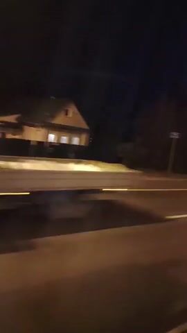 The video shows a nighttime view from a moving vehicle. The scene is dimly lit, with streetlights casting a warm glow on the asphalt road. Trees and a building with illuminated windows are visible in the background. The vehicle appears to be traveling along a highway, as indicated by the white lines marking the lanes. A truck with advertisements on its side is seen driving past in the opposite direction. The overall impression is of a typical nighttime drive on a road with some commercial or industrial development.