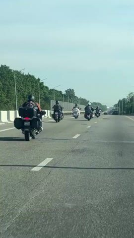 The video shows a group of motorcycles riding on a highway. The motorcycles are in a staggered formation, with several riders in the foreground and more further down the road. The weather appears to be clear and sunny. The highway has multiple lanes and is bordered by trees and a guardrail. The perspective suggests the video is being recorded from another vehicle on the highway.