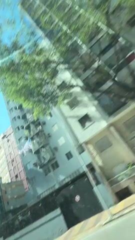 The video shows a low-angle view of several tall buildings under a clear blue sky, as if seen from a moving vehicle. The camera pans from left to right, revealing different facades and architectural styles. The buildings are mostly residential or commercial, with visible balconies, windows, and some air conditioning units. Palm trees and other foliage can be seen occasionally, adding greenery to the urban landscape. The sunlight is bright, creating strong shadows and reflections on the glass surfaces. The movement of the camera suggests a dynamic perspective, possibly from a car driving through a city street.