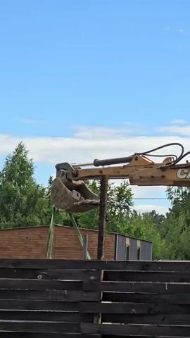 An excavator is lifting a large metal pole out of the ground. The excavator is yellow with the brand name "CASE" visible on the arm. The pole is dark brown and appears to be made of wood or metal. The excavator is positioned behind a dark wooden fence. In the background, there are green trees, a wooden building, and a blue sky with scattered clouds. The overall scene suggests a construction or demolition site.