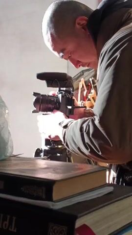 A bald man is standing, carefully adjusting a camera mounted on a tripod. The camera has a microphone attached. He is looking at the camera. The camera and tripod are set on a stack of books. There is a plastic bag next to the books. Sunlight is coming from behind the camera, illuminating the man and the equipment. The wall behind the man is white. He is wearing a brown jacket.